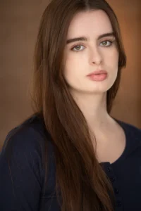 Professional female actor posing for a studio headshot with natural makeup and a dark outfit against a neutral background.